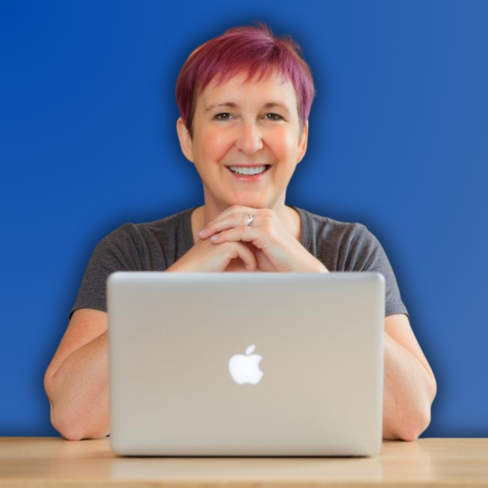 Woman with very short deep pink hair sitting at her laptop, smiling at the canera
